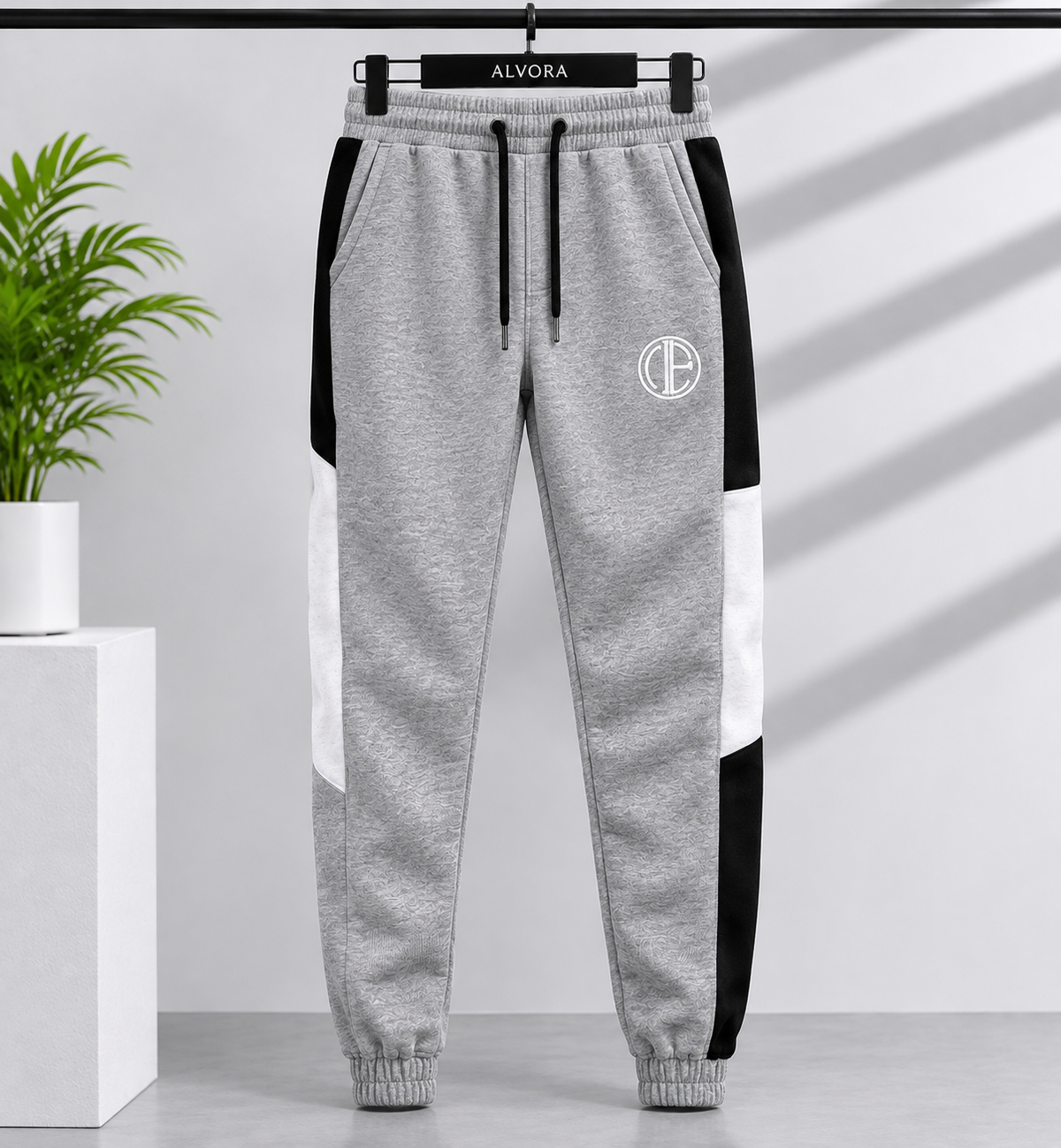 Gray heather jogger sweatpants with black and white side panels, a black drawstring, and a circular white monogram logo on the left thigh, hanging on a black hanger labeled ALVORA against a light gray wall with shadow patterns.
