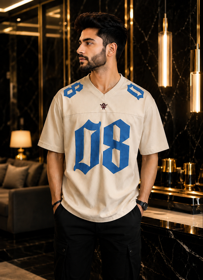 A man wearing a cream-colored football-style jersey with bold blue numbers '08' on the front and blue shoulder details, standing in a luxurious interior with dark marble walls and gold pendant lights.