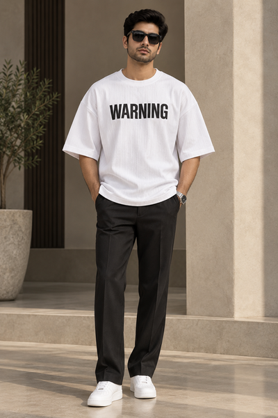 A man wearing a white short-sleeve t-shirt with 'WARNING' printed in black across the chest, paired with black trousers and white sneakers, standing in a modern architectural setting.
