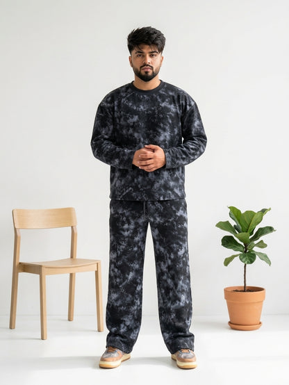 A man wearing a matching dark gray and black tie-dye sweatshirt and sweatpants set, standing in a studio with a wooden chair and a potted plant.