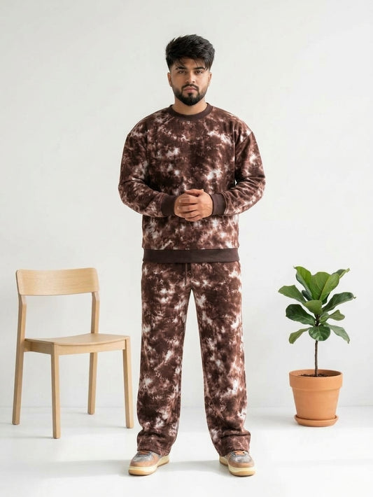A man wearing a matching brown and white tie-dye sweatshirt and sweatpants set, standing in a studio with a wooden chair and a potted plant.