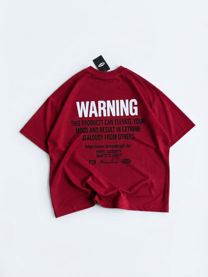A dark red short-sleeve t-shirt laid flat on a light gray background, showing the back side with a large white 'WARNING' text graphic and smaller black text below.
