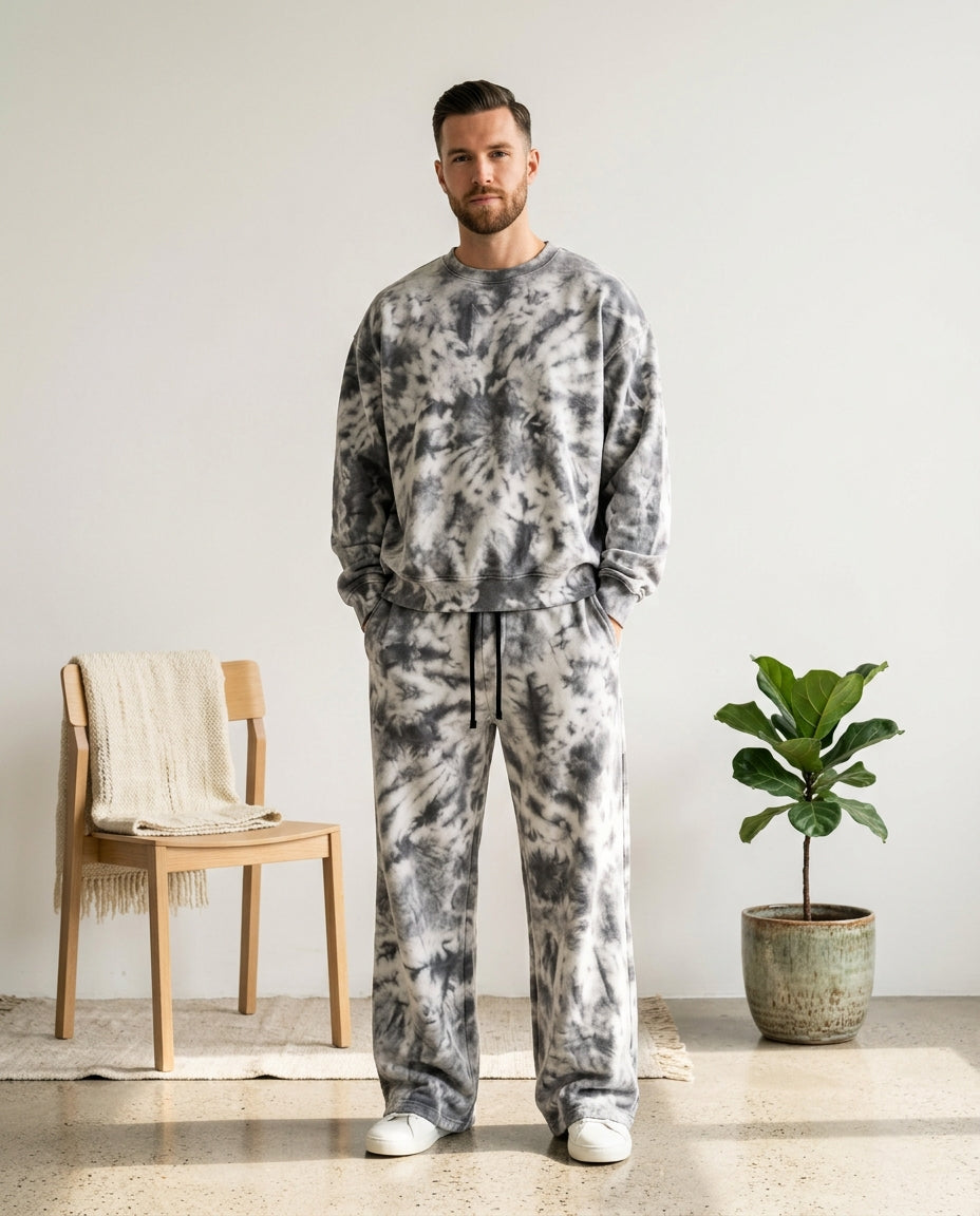 A man wearing a matching gray and white tie-dye sweatshirt and sweatpants set, standing in a minimalist room with a wooden chair and a potted plant.