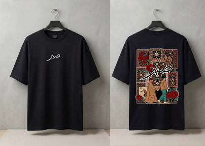 A black short-sleeve T-shirt shown from both front and back on hangers against a light gray wall. The front has a small white script graphic on the left chest, and the back features a large rectangular multicolor graphic with floral and script artwork.