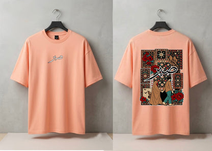 A peach-colored short-sleeve t-shirt shown from both front and back views, hanging on black hangers against a textured gray wall. The front features a small white Arabic calligraphy graphic on the left chest, while the back displays a large, detailed graphic print with Arabic calligraphy, a woman in a hijab, and a traditional patterned rug design.