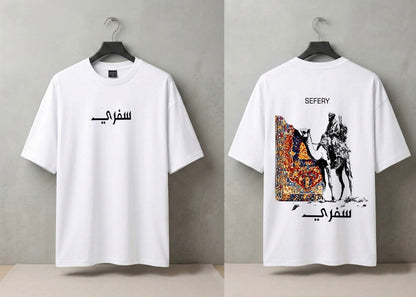 A white short-sleeve T-shirt shown from both front and back on black hangers against a gray wall. The front has black Arabic text across the chest, and the back has a large graphic print featuring a camel, rider, and ornate patterned artwork, with 'SEFERY' above and Arabic text below.