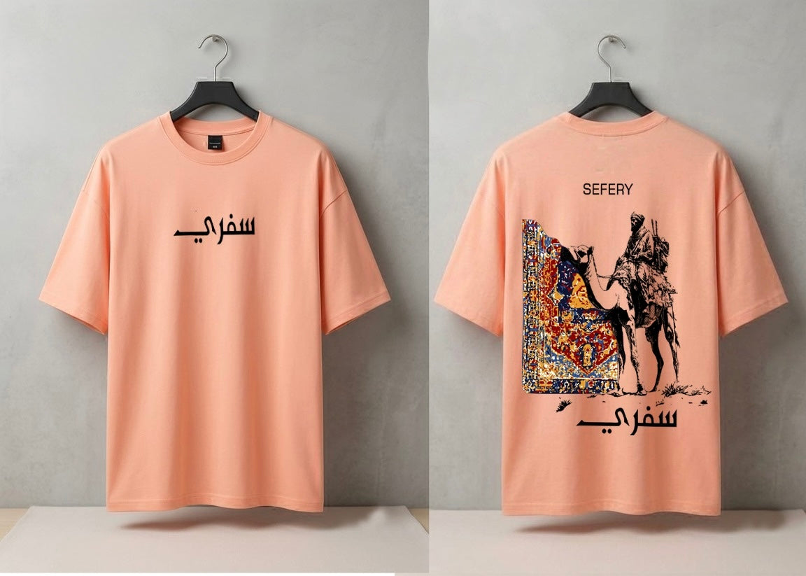 A peach-colored t-shirt shown from both front and back views, hanging on black hangers against a gray wall. The front features black Arabic calligraphy, while the back displays a large graphic print of a camel and rider with a colorful patterned rug, alongside the text 'SEFERY' and Arabic calligraphy.