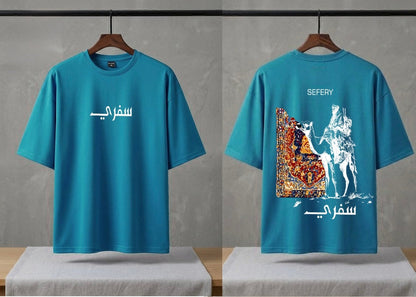 A teal blue short-sleeve t-shirt shown from both front and back on wooden hangers. The front features white Arabic calligraphy, while the back displays a large graphic print of a white horse and rider with a colorful patterned rug, alongside white English text reading 'SEFERY' and Arabic calligraphy.