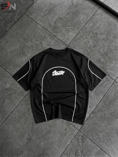 A black short-sleeve t-shirt with white contrast piping and a white graphic logo on the chest, laid flat on a dark gray tiled floor.