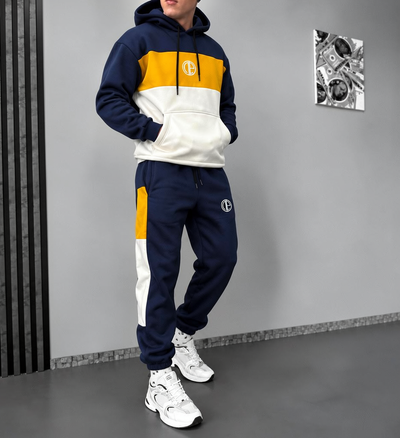 A man modeling a matching two-piece tracksuit with a color-blocked hoodie and jogger pants. The hoodie features navy blue, yellow, and off-white panels, with a circular 'TC' logo on the chest and thigh.