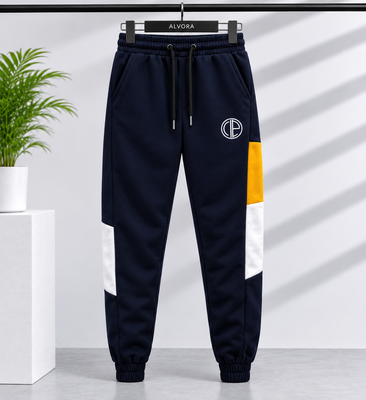 Navy blue jogger sweatpants with white and yellow color-block panels on the legs, featuring a white circular logo on the left thigh. The pants are hanging on a black hanger labeled 'ALVORA' against a light gray wall with diagonal shadows.