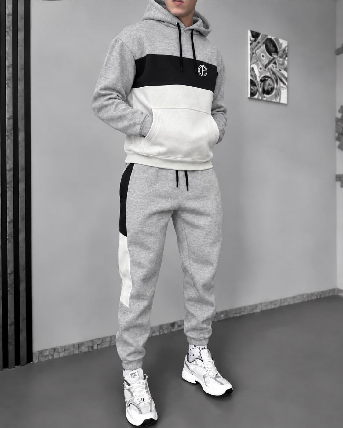 A person modeling a matching two-piece tracksuit in a gray indoor setting. The hoodie has a color-blocked design with gray, black, and white sections, and the jogger pants have gray with black and white side panels.