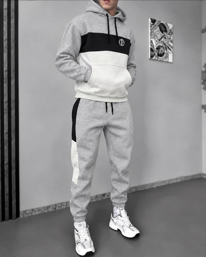 A person modeling a matching two-piece tracksuit in a gray indoor setting. The hoodie has a color-blocked design with gray, black, and white sections, and the jogger pants have gray with black and white side panels.